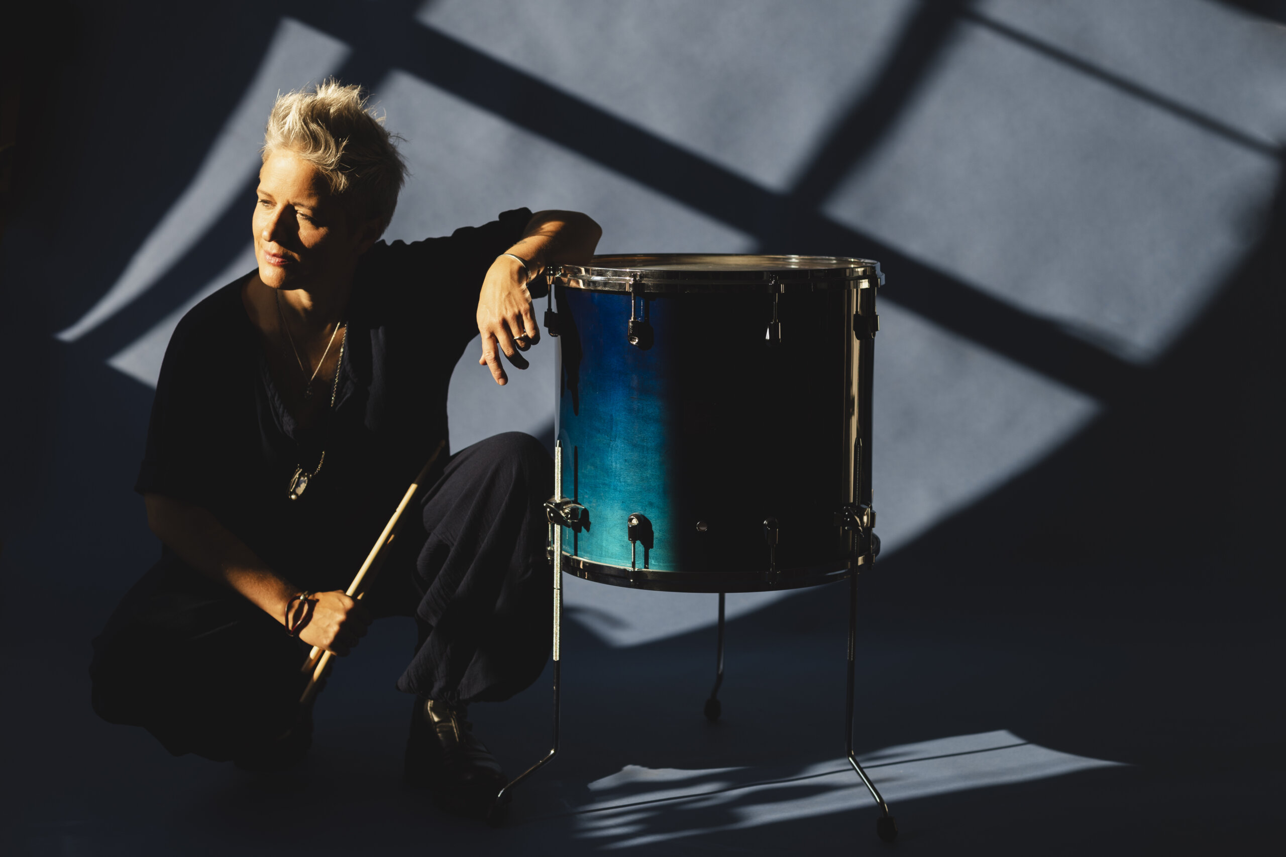 A person with short gray hair and tattoos, wearing a sleeveless black shirt, sits on rocks by a body of water, smiling and looking to the side with a bridge visible in the background—reminiscent of jazz band leader Allison Miller of Boom Tic Boom.
