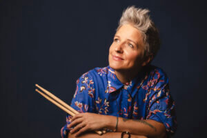 Smiling person with short, spiky gray hair holds drumsticks and looks upward, channeling the vibrant energy of Allison Miller’s Boom Tic Boom. They wear a blue, floral-patterned shirt and have tattoos on their forearm. Dark, plain background.