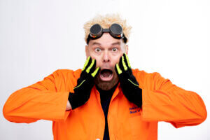 A man with spiky blond hair and goggles on his head wears an orange lab coat and black gloves with neon green stripes, holding his face in surprise with his mouth open wide.