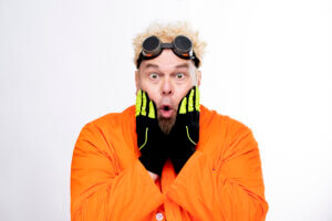 A surprised man with spiky blond hair, wearing black goggles on his head, black gloves with yellow stripes, and an orange lab coat, holding his face with both hands against a white background.