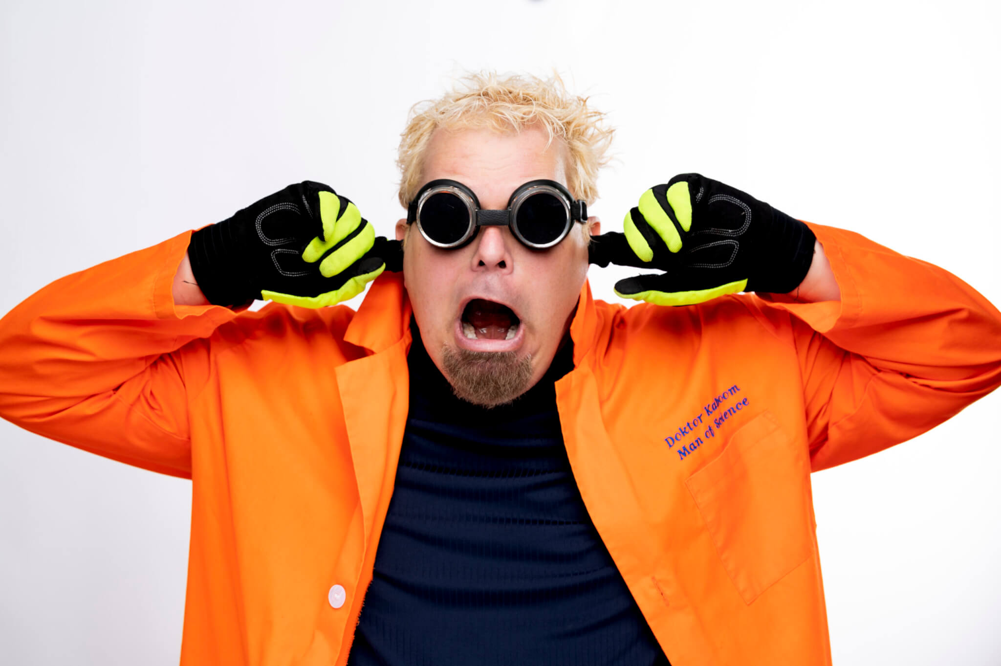 A man with spiky blond hair and a goatee wears an orange lab coat, black gloves with yellow accents, and round black goggles. He covers his ears with his fingers and makes a surprised, wide-mouthed expression.