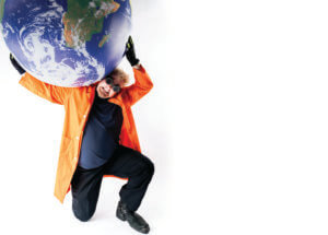 A person in an orange coat, black gloves, and goggles kneels while humorously holding up a large globe above their shoulders against a white background, mimicking the pose of Atlas.