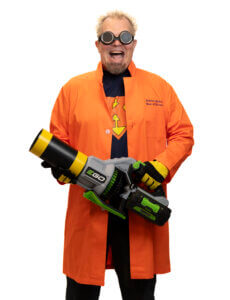 A man with spiky blond hair wearing goggles, an orange lab coat, yellow gloves, and a dark shirt holds a large green and gray leaf blower, smiling excitedly at the camera.