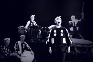A group of performers in traditional Japanese attire play taiko drums on stage. One person stands at the front singing or speaking, while others play drums in the background. The scene is lit dramatically.