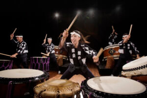 Five performers in traditional attire enthusiastically play Japanese taiko drums on stage, each holding drumsticks mid-motion, with energetic expressions and dramatic lighting highlighting the scene. From KODO: Luminance