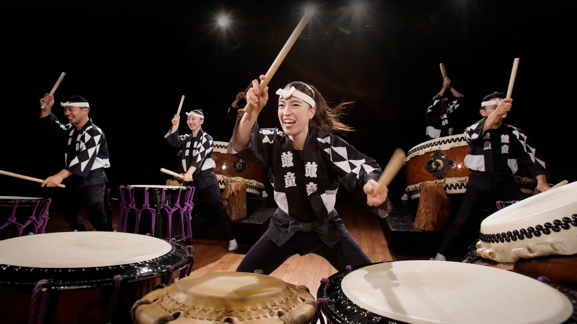 A group of performers in traditional attire energetically play Japanese taiko drums on a wooden stage, smiling and moving in unison under stage lights at the Purdue Convocations 2026–27 season unveiling.