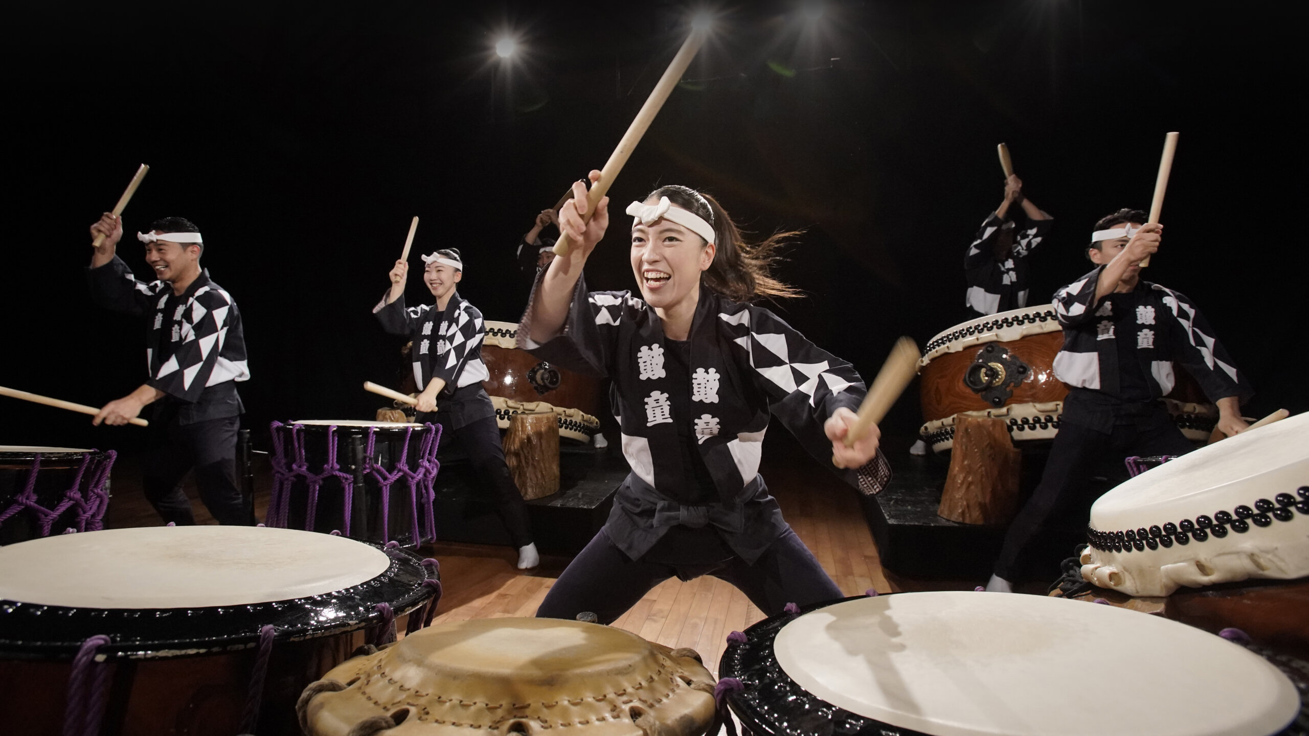 A group of performers in traditional attire energetically play Japanese taiko drums on a wooden stage, smiling and moving in unison under stage lights at the Purdue Convocations 2026–27 season unveiling.