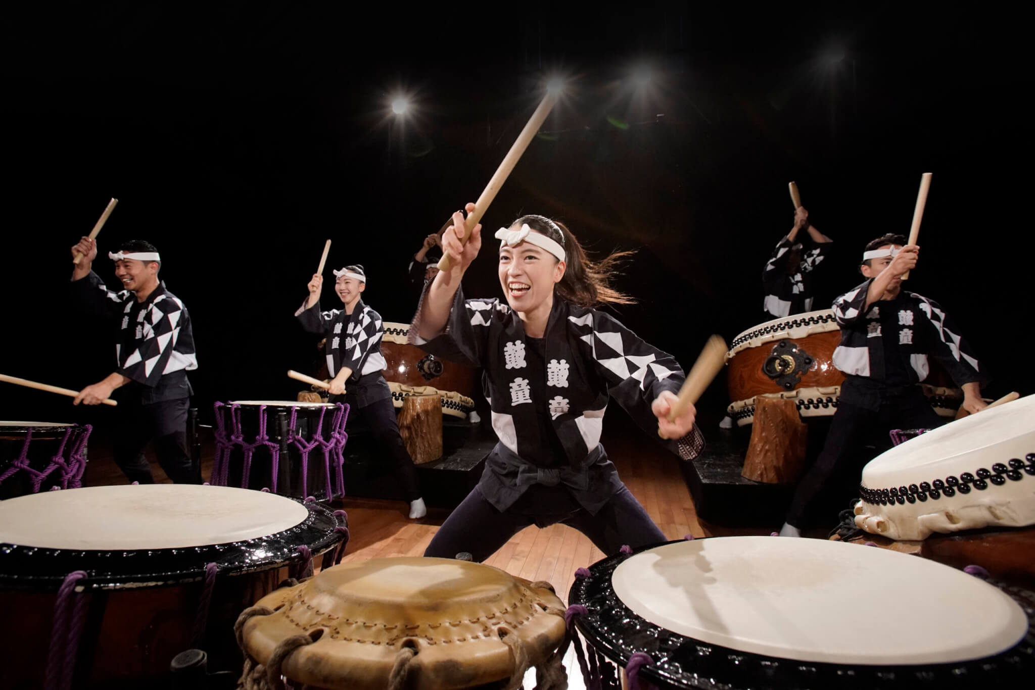 Five performers in traditional attire enthusiastically play Japanese taiko drums on stage, each holding drumsticks mid-motion, with energetic expressions and dramatic lighting highlighting the scene. From KODO: Luminance