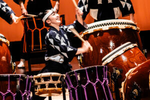 A smiling performer in traditional Japanese attire energetically plays large taiko drums on stage, surrounded by other drummers and drums, against an orange backdrop.
