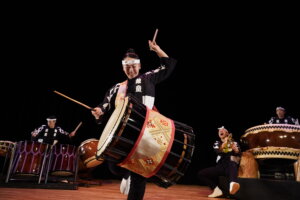 A group of Japanese taiko drummers perform energetically on stage, wearing traditional black and white costumes. The central drummer smiles and lifts a drumstick in the air, while others play drums in the background.