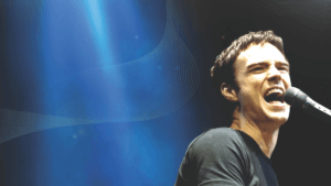 A man in a black shirt passionately sings into a microphone against a dramatic blue background, embodying the energy of The Music of Billy Joel and Elton John starring Michael Cavanaugh, with light rays and abstract wave patterns shining behind him.