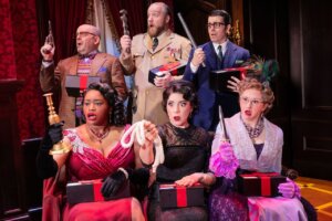 Six people in colorful, vintage costumes sit or stand close together, each holding a different object and a gift box, looking surprised or alarmed—evoking the playful flair of Kinky Boots. Ornate red curtains and a dark wooden wall set the scene.