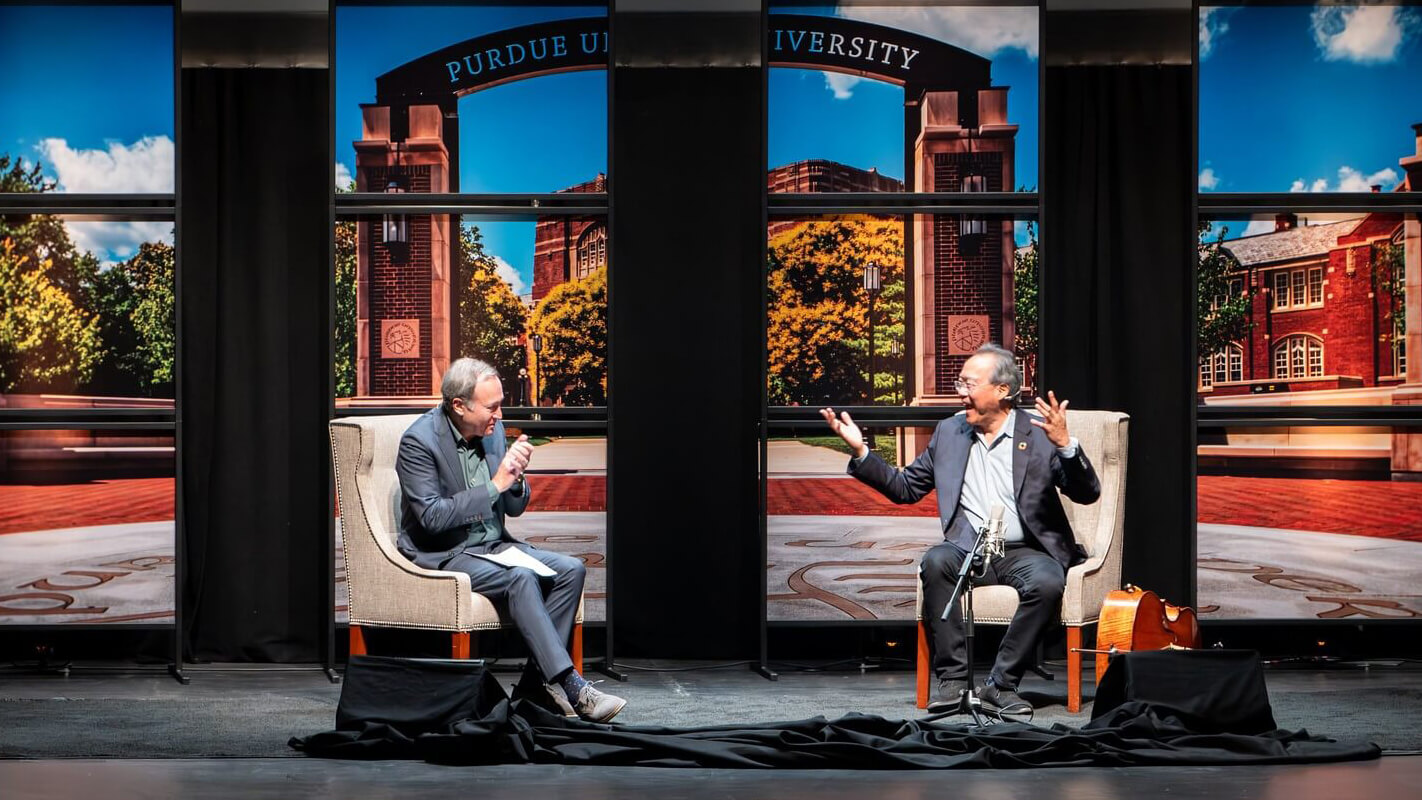 Jeffrey Brown from PBS NewsHour and cellist and humanitarian Yo-Yo Ma sit on stage in chairs having a discussion, microphones in hand. Large panels display scenic images of Purdue University, highlighting Purdue Convocations. The stage is simply decorated, with cords and a small table between them.