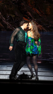Two dancers in vibrant green and blue costumes perform a dramatic, close pose on stage, appearing to be mid-dance, with theatrical lighting and a dark, nature-themed backdrop behind them.