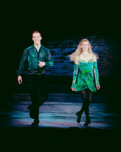 Two dancers in green costumes perform an Irish dance on stage, smiling as they step in sync. The background is dark with subtle lighting highlighting the dancers.