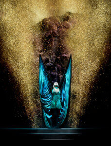 A performer in a green costume stands on stage with large, dramatic wings spread wide, surrounded by a burst of golden glitter or smoke against a dark background.