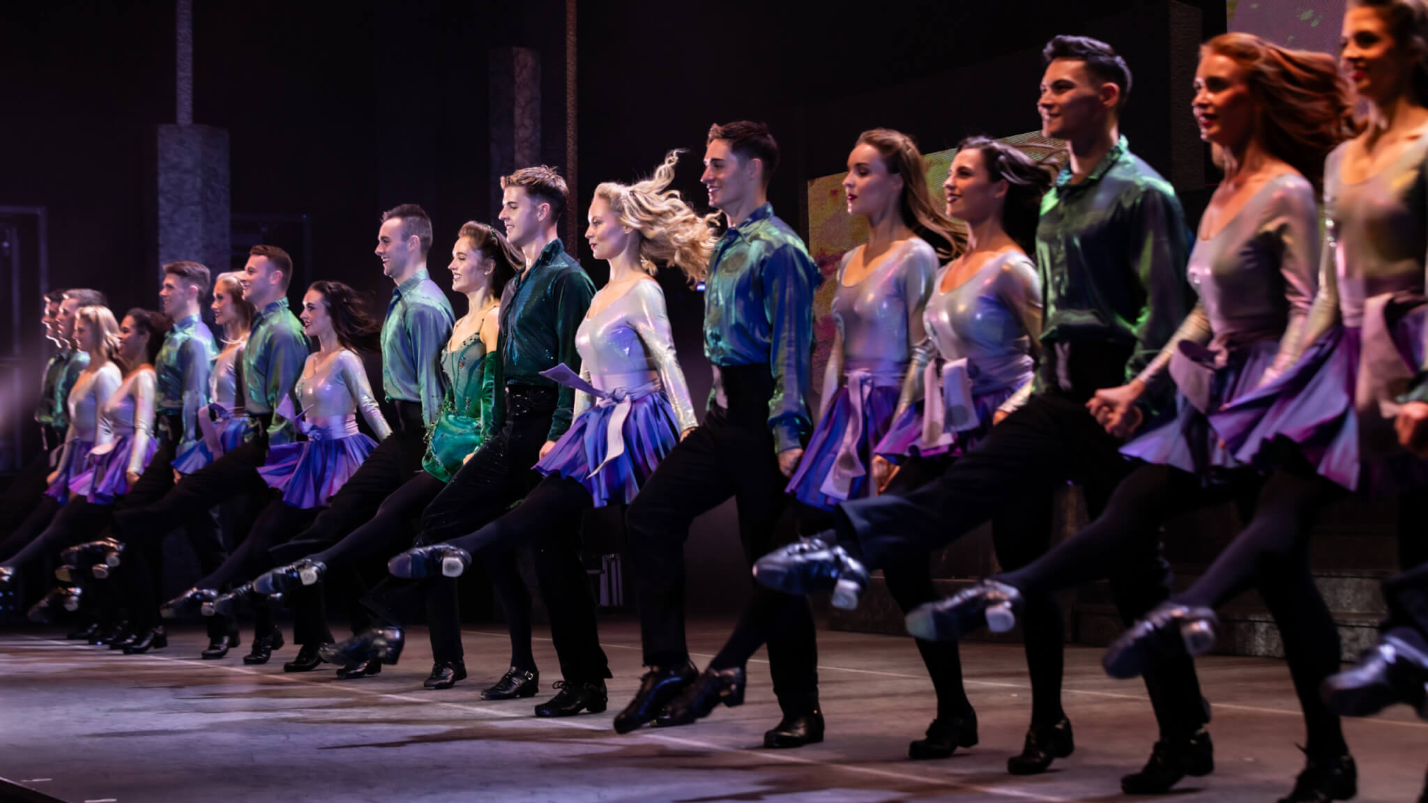 A line of dancers in coordinating costumes performs a synchronized high-kick routine on stage, with motion and energy evident in their movements and expressions.