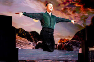 A male dancer in a green shirt and black pants leaps gracefully with arms extended, performing on stage with a scenic backdrop of mountains, water, and a dramatic, colorful sky.