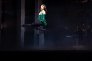 A female dancer in a green costume and black tights performs a high leap on stage, with dramatic lighting highlighting her in midair and musicians playing in the dimly lit background.