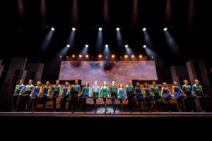A large group of performers in colorful costumes dances in a straight line on stage, with dramatic lighting and a vivid backdrop displaying a cloudy sky.