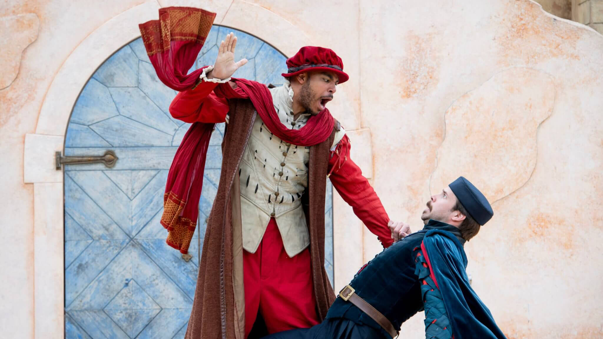 Two actors in colorful, Renaissance-style costumes perform a dramatic scene as The Lord Chamberlain’s Men present William Shakespeare’s OTHELLO; one stands, raising his hand, while the other is on the ground, clutching the standing actor’s robe against a textured wall and a blue wooden door.