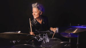 A smiling person with short, gray hair plays a drum set, holding drumsticks. Channeling the energy of Allison Miller’s Boom Tic Boom, they wear a black shirt and are lit dramatically against a dark background, with light trails visible above the drums.