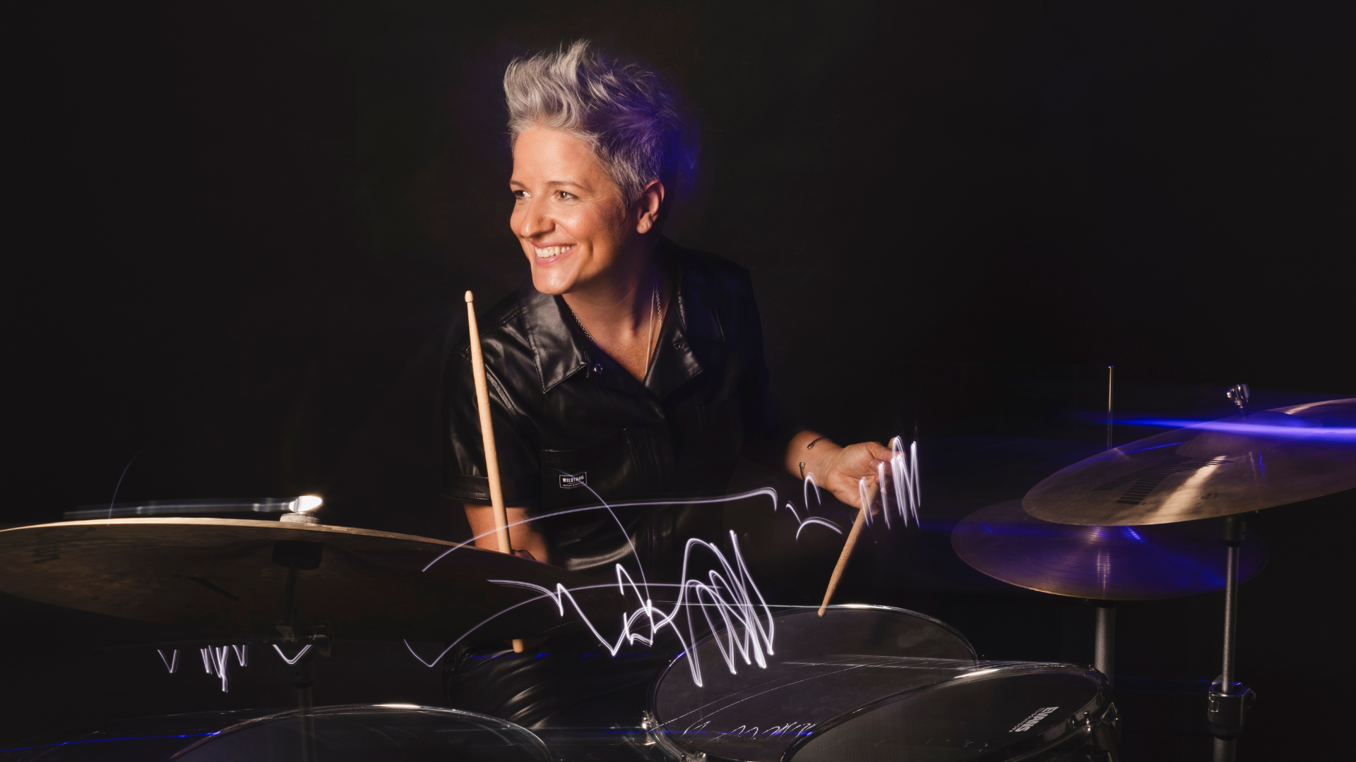 A smiling person with short, gray hair plays a drum set, holding drumsticks. Channeling the energy of Allison Miller’s Boom Tic Boom, they wear a black shirt and are lit dramatically against a dark background, with light trails visible above the drums.