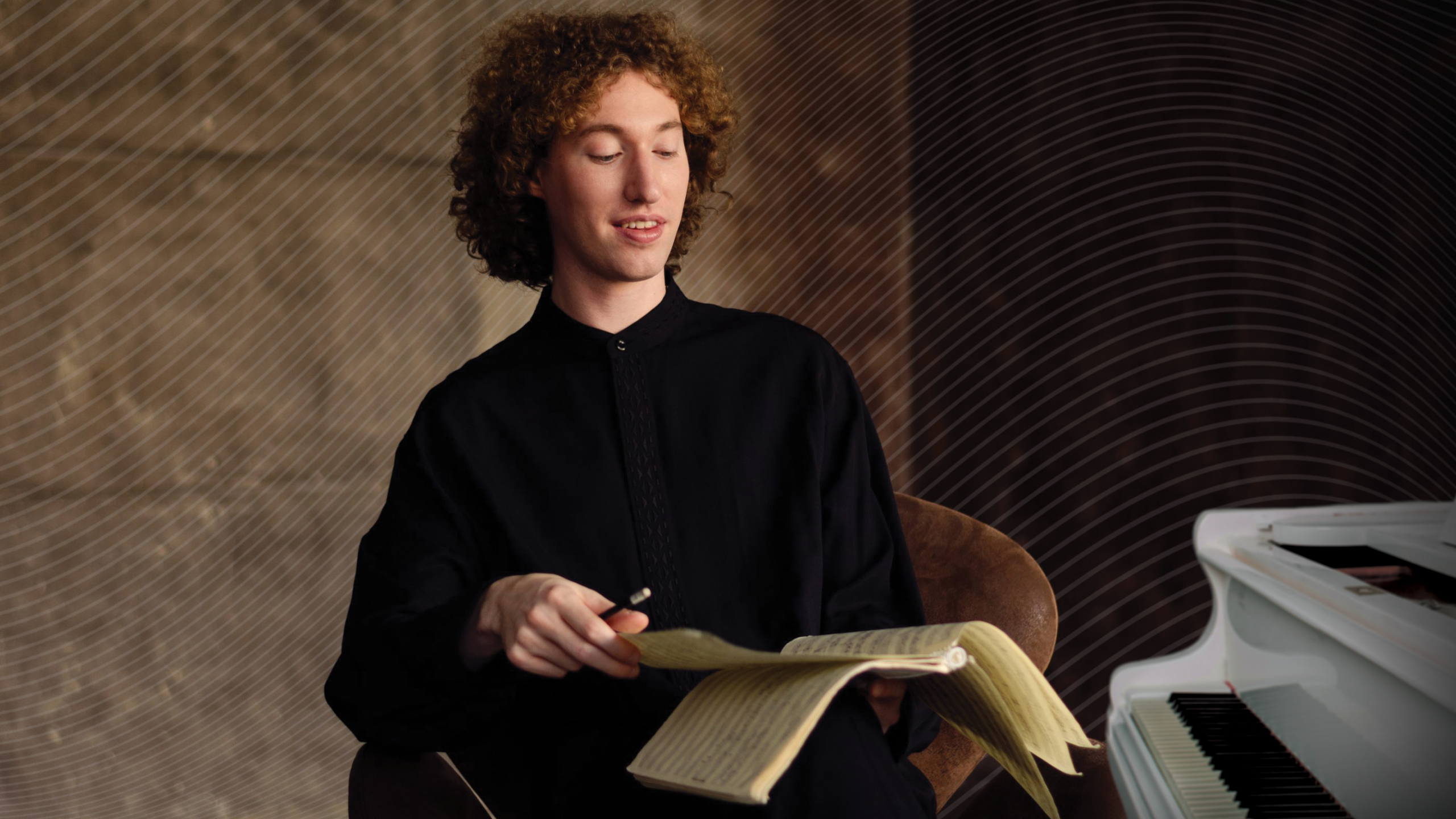 Pianist Michael Davidman against a brown, patterned background. They have curly hair, is wearing a black shirt, and sits in a chair, holding sheet music and a pencil, with part of a white piano visible beside them.