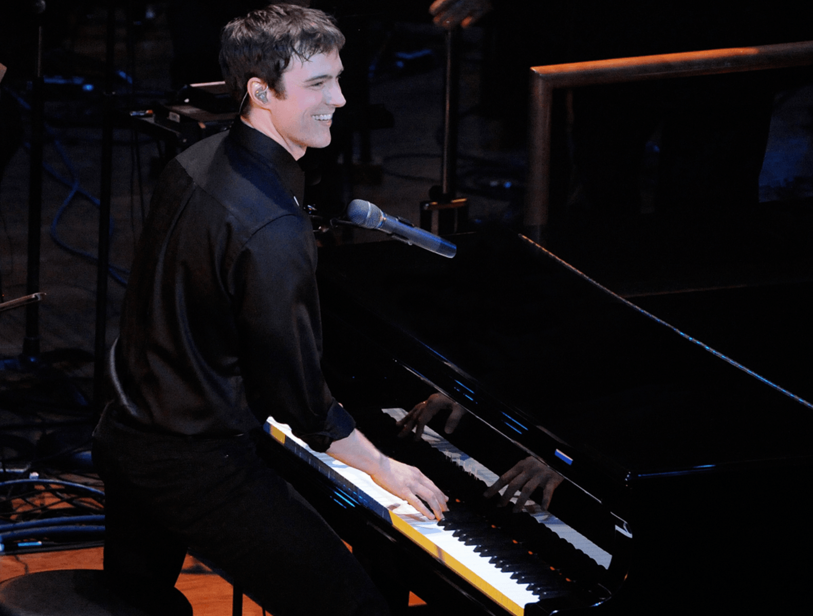 Michael Cavanaugh, smiling, wearing a black shirt plays a grand piano on stage with a microphone nearby. Stage equipment and cables are visible in the background.