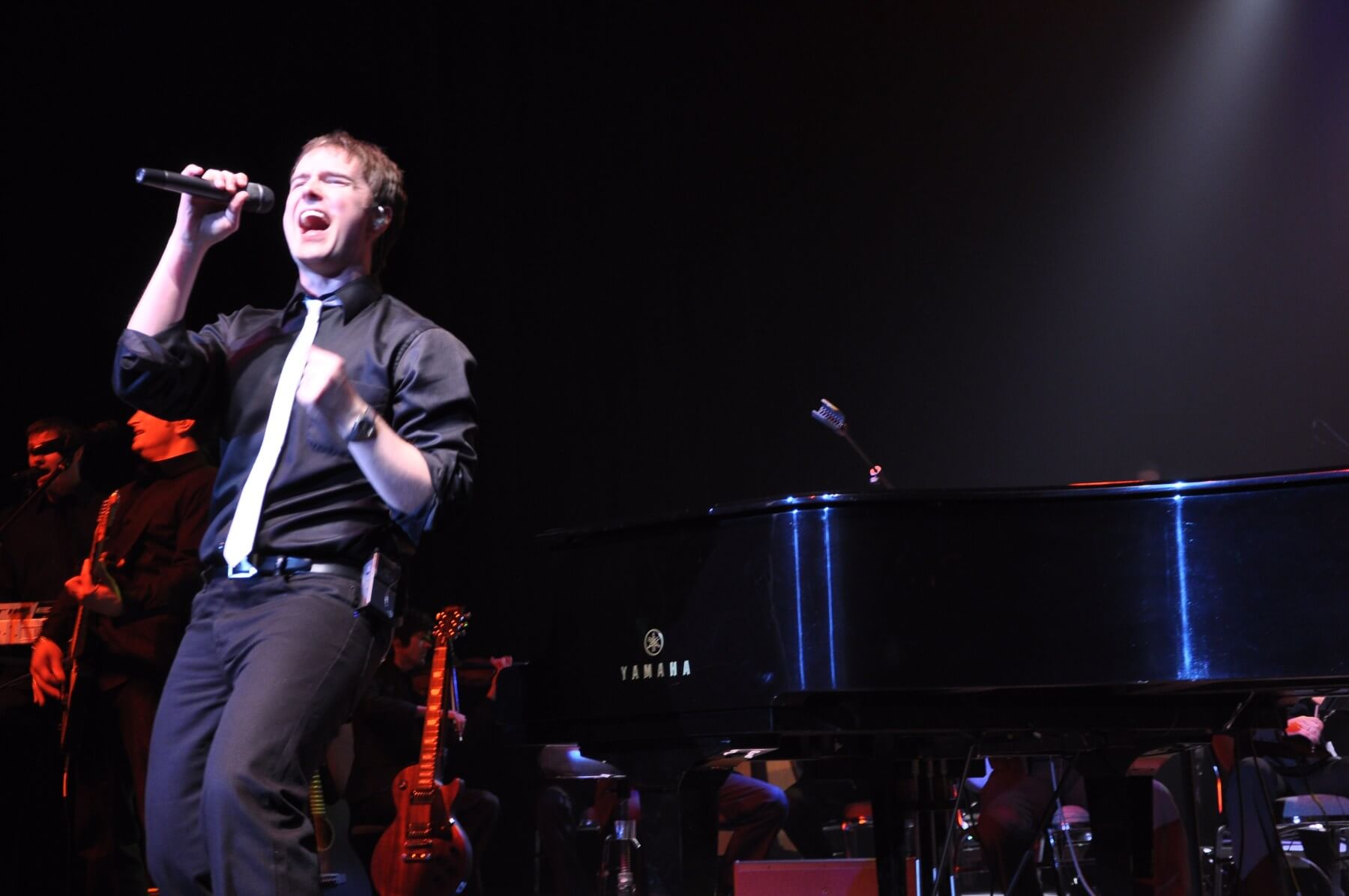 A smiling person in a black shirt performs on a piano on stage, reminiscent of Billy Joel, with a microphone positioned nearby and stage equipment visible in the background.