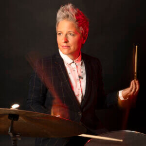 A person with short, spiked gray hair wearing a suit plays the drums, holding drumsticks. The lighting is dramatic, with red highlights on their face and suit, set against a dark background.