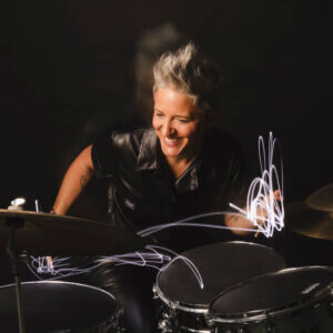A smiling person with short, spiky gray hair plays drums, wearing a black leather shirt. Light trails swirl around their drumsticks, emphasizing motion against a dark background.