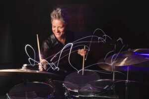 A smiling person with short gray hair plays the drums, creating swirling light trails in the air, against a dark background.