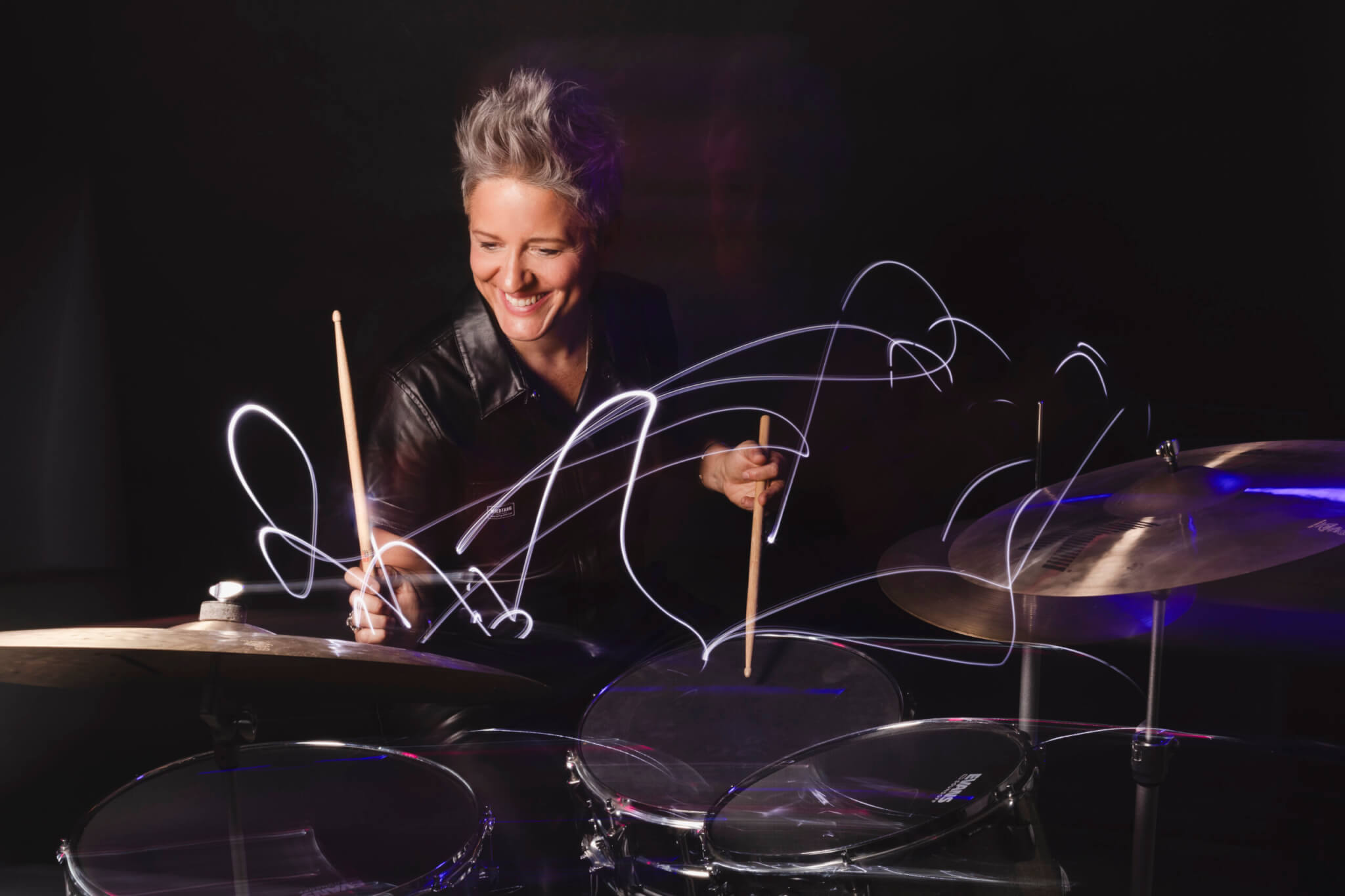 A smiling person with short gray hair plays the drums, creating swirling light trails in the air, against a dark background.