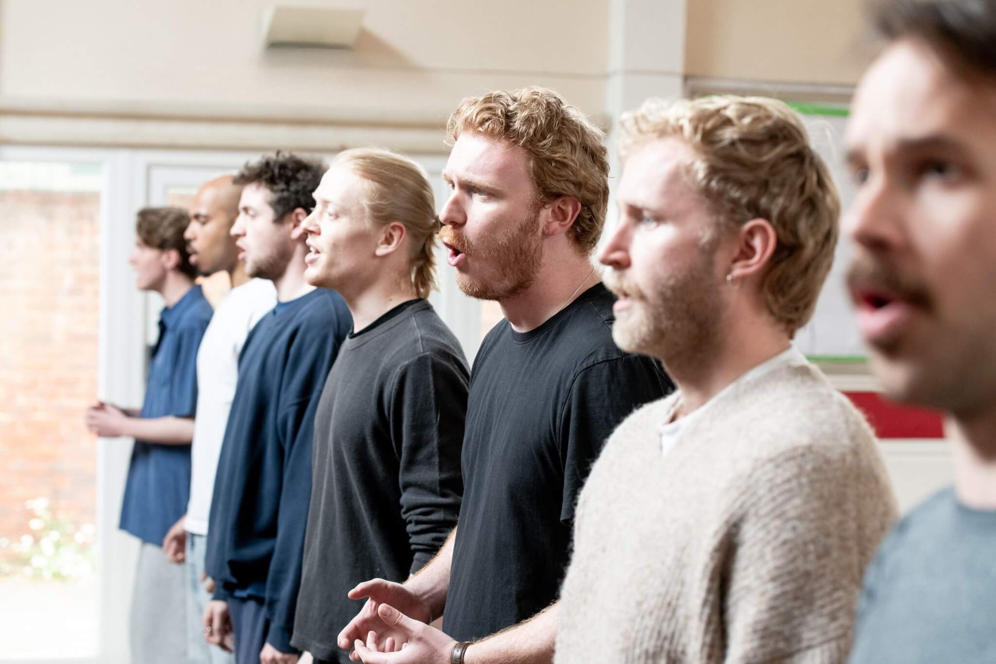 A group of seven men stand in a row indoors, singing together with focused expressions. Dressed casually as sunlight streams in from a nearby window, they rehearse for an upcoming event—hoping their audience will RSVP soon.