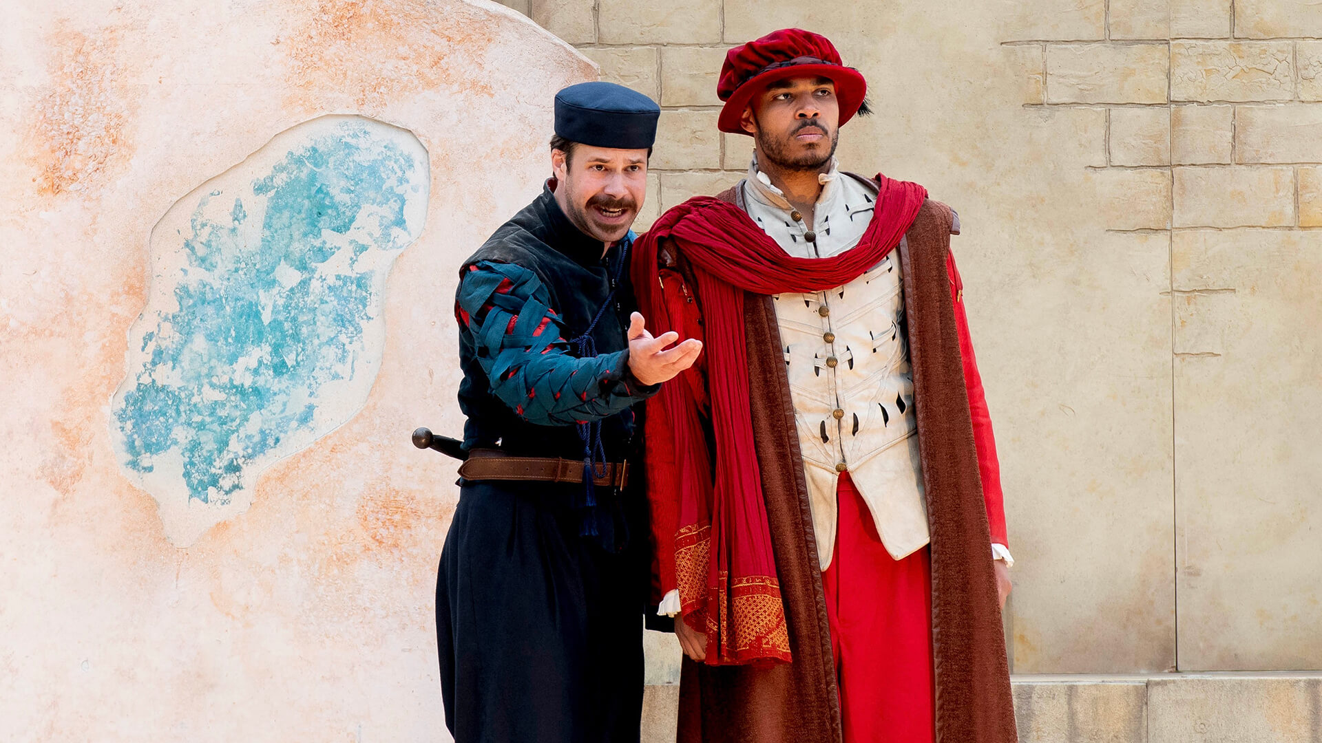Two actors in colorful period costumes perform on stage outdoors, delivering a dynamic introduction to Shakespeare’s Othello: one gestures passionately while the other looks serious, standing near a textured wall with a large, blue-stained area.
