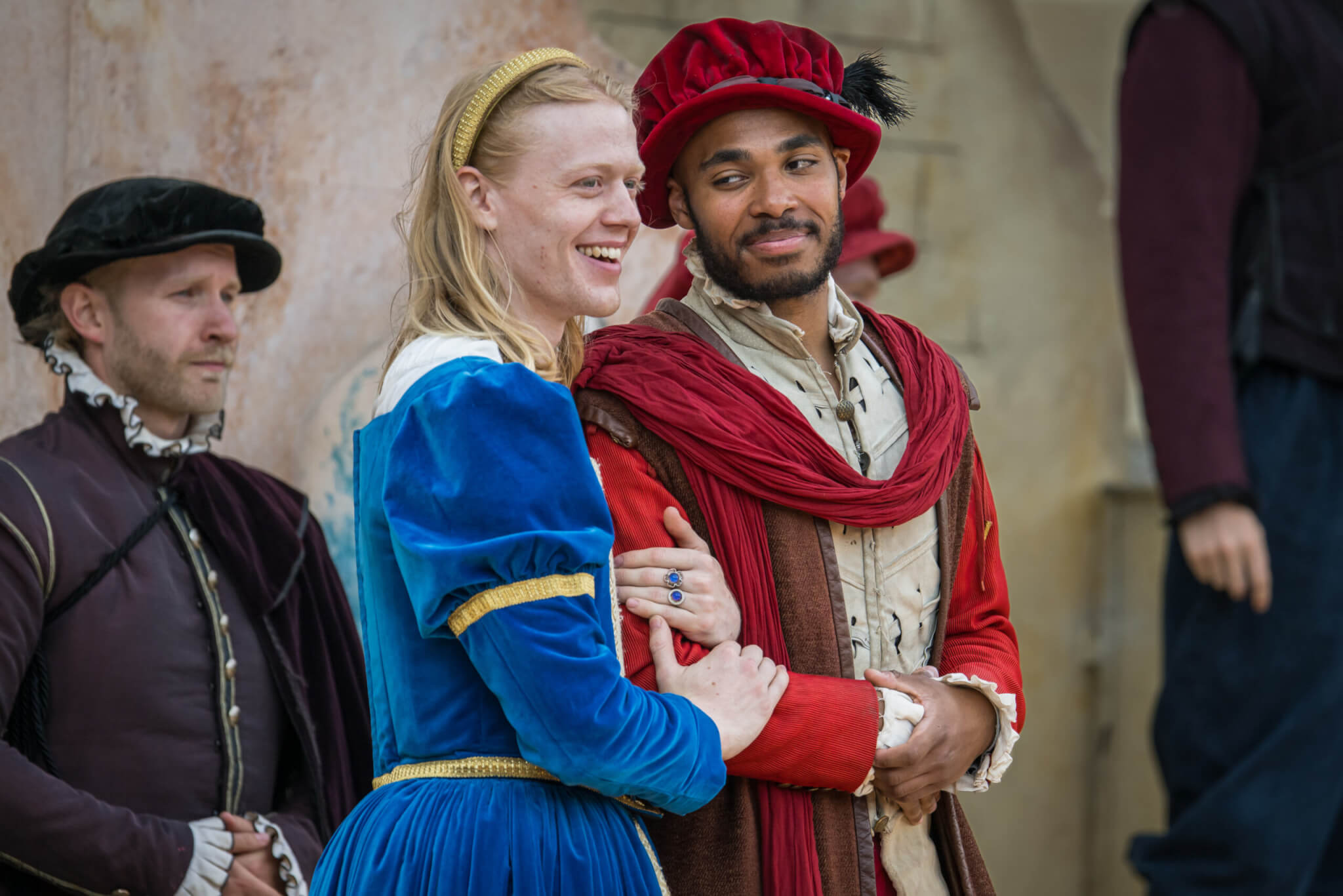 cast of Othello: two people in colorful Renaissance costumes stand close together, smiling. One wears a blue dress and yellow headband; the other wears red and white with a red cap. Another person in dark clothing stands in the background, adding to the festive atmosphere.
