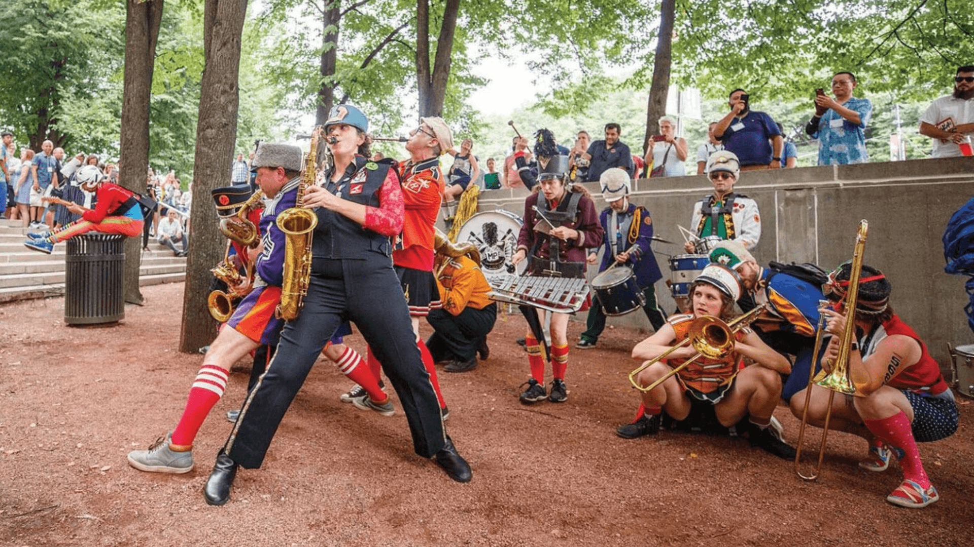 A lively street band in colorful, eccentric costumes performs energetically in a park during one of West Lafayette’s vibrant musical performances, as a crowd watches. Musicians play brass and percussion instruments under green trees on a dirt path, creating an atmosphere reminiscent of Purdue Convocations events.