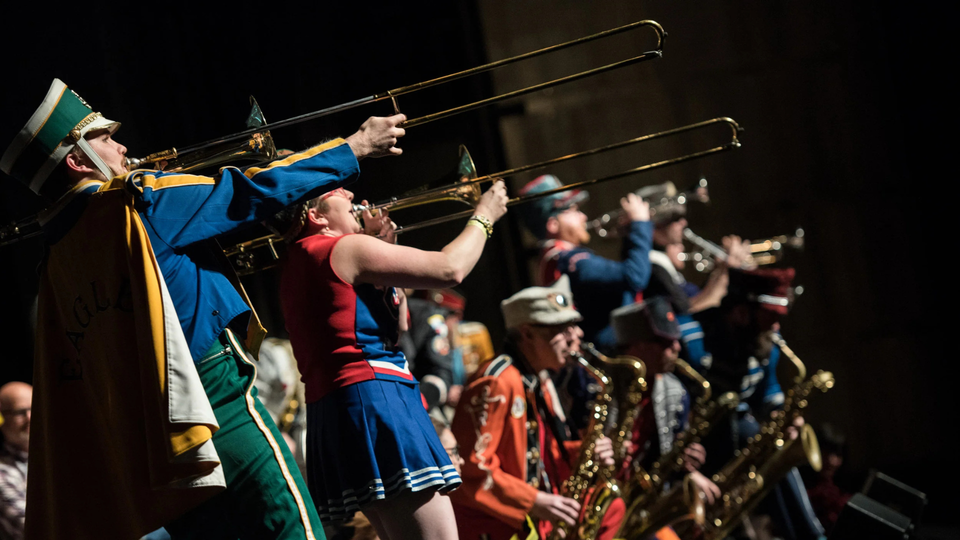 A marching band in colorful uniforms plays brass instruments energetically on stage, with trombone and saxophone players leaning forward in unison during one of the free musical performances presented by Purdue Convocations in West Lafayette.