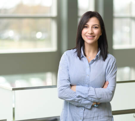 Tulin Tosun, picutired here with dark hair and a light blue shirt stands indoors with her arms crossed, smiling, in front of large windows with blurred outdoor scenery.