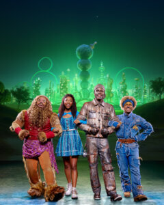 Four people in colorful, whimsical costumes stand smiling in front of a glowing, futuristic green city under a starry night sky.