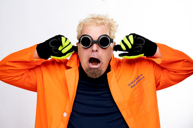 A man in an orange lab coat and gloves covers his ears, wearing round black goggles and making a surprised face against a plain white background.