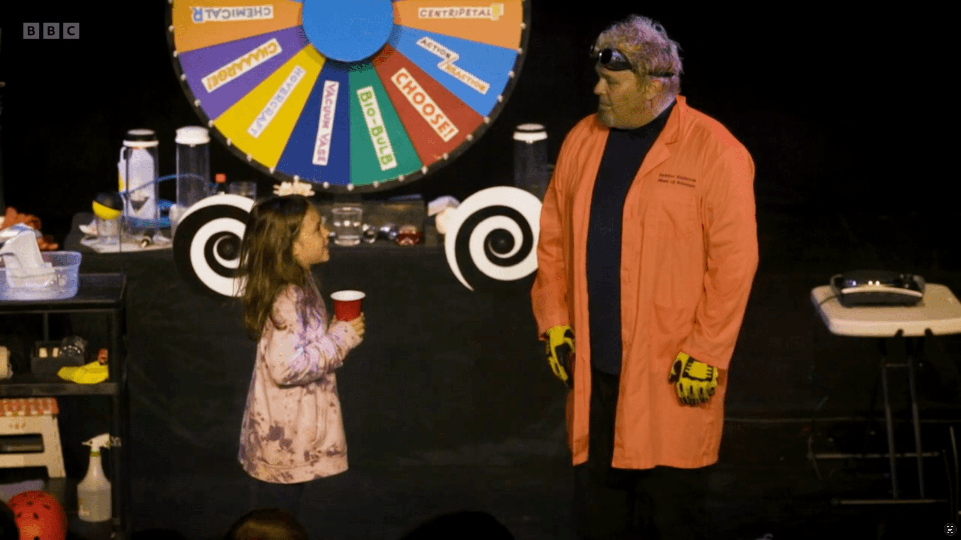 Doktor Kaboom (David Epley) in an orange lab coat and goggles stands on stage beside a young girl holding a red cup, with a colorful prize wheel and science equipment in the background, exemplifying Technical Educational Outreach during Purdue Convocations.