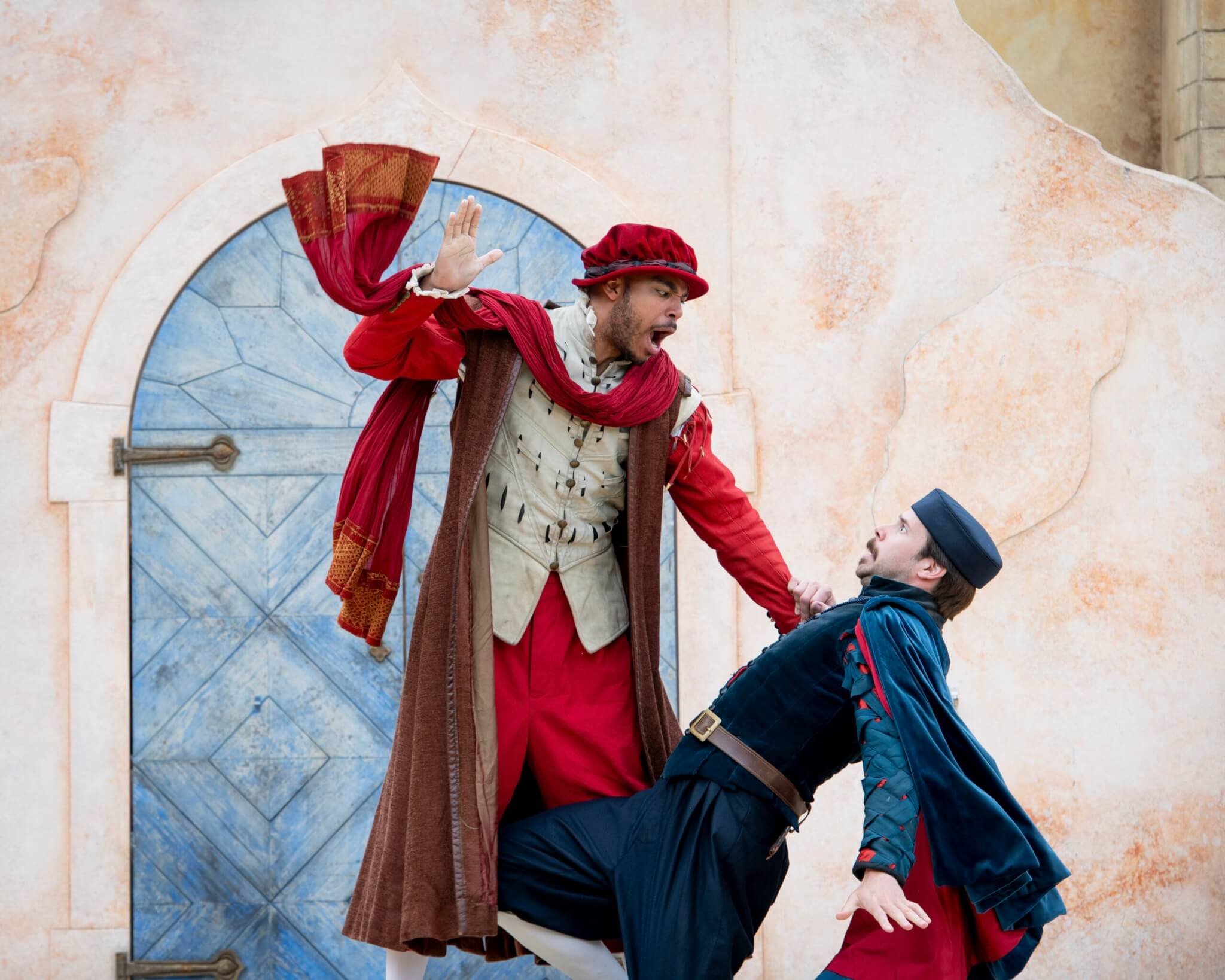 Two men in colorful Renaissance costumes act out a dramatic scene; one raises his hand as if to strike, while the other leans back in surprise or fear in front of a weathered wooden door.