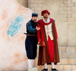 Two actors in period costumes perform on an outdoor stage. One wears a dark outfit and gestures expressively, while the other wears red and brown robes, standing still beside him against a stone-like backdrop.