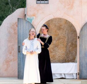 Two actors in period costumes perform outside; one in a white dress looks emotional, and another in a black dress stands behind, comforting her. An arched doorway and bed are visible in the background.
