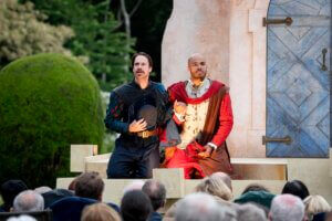 Two actors in period costumes perform dramatically on an outdoor stage before an audience. One holds a hat to his chest; the other wears red and looks upward. A faux stone wall and blue door are behind them.