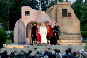 Seven actors in period costumes stand on stage in front of a rustic, beige set resembling old buildings, while an outdoor audience watches. Lush green trees are visible in the background.
