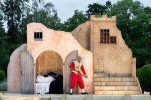 An actor in a red costume performs on an outdoor stage set resembling a stone building, with one person lying in a bed and trees visible in the background.
