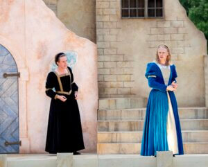 Two people in Renaissance-style dresses perform on an outdoor stage with stone steps and a painted backdrop, suggesting a historical or Shakespearean play.